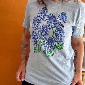 May include: A light blue t-shirt featuring a graphic of bluebonnet flowers in shades of blue and green. The floral design is centered on the front of the shirt. The shirt has short sleeves and a crew neck.