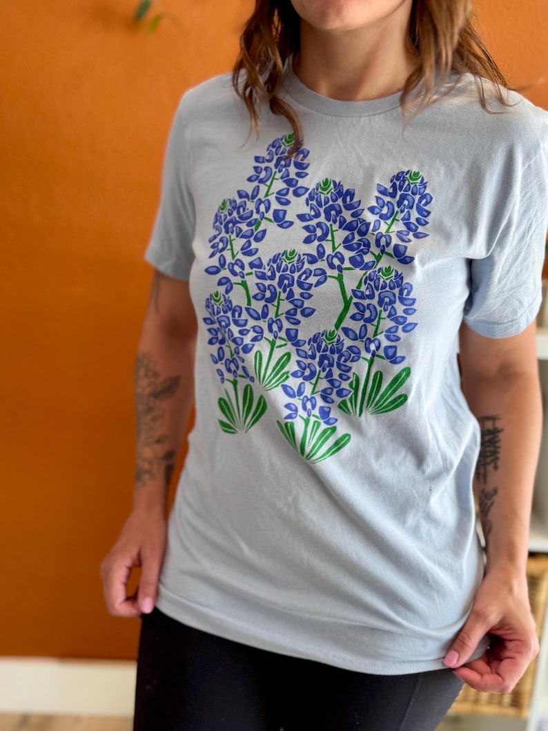 May include: A light blue t-shirt featuring a graphic of bluebonnet flowers in shades of blue and green. The floral design is centered on the front of the shirt. The shirt has short sleeves and a crew neck.