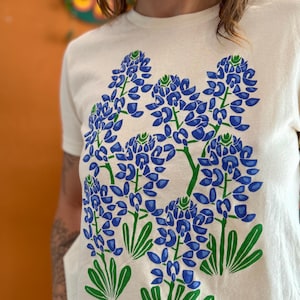 May include: Cream-colored t-shirt featuring a large graphic of bluebonnet flowers in shades of blue and green. The design includes detailed illustrations of the flowers and leaves, creating a botanical theme.