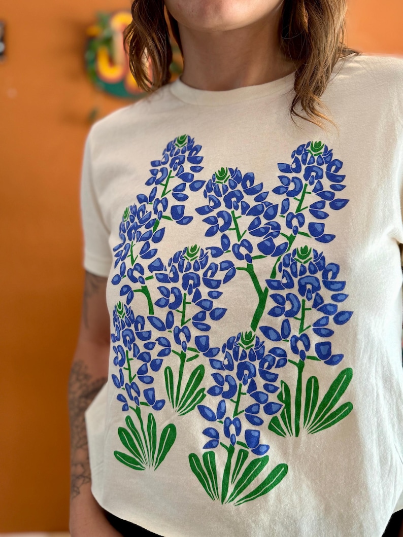 May include: Cream-colored t-shirt featuring a large graphic of bluebonnet flowers in shades of blue and green. The design includes detailed illustrations of the flowers and leaves, creating a botanical theme.