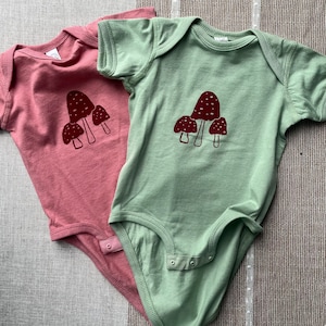 May include: Two baby onesies with short sleeves. One is pink with a mushroom graphic and the other is green with a mushroom graphic.