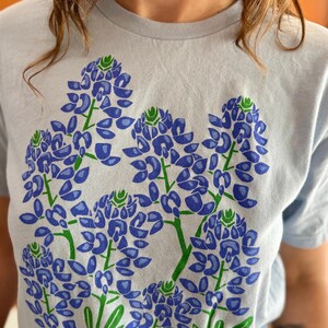 May include: Light blue t-shirt featuring a large floral design of bluebonnets. The graphic print showcases detailed blue and green flowers with green stems and leaves. The shirt is a soft, casual style.