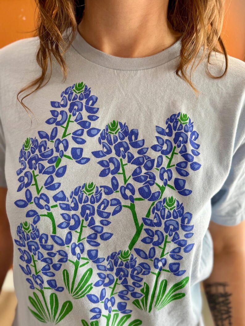 May include: Light blue t-shirt featuring a large floral design of bluebonnets. The graphic print showcases detailed blue and green flowers with green stems and leaves. The shirt is a soft, casual style.