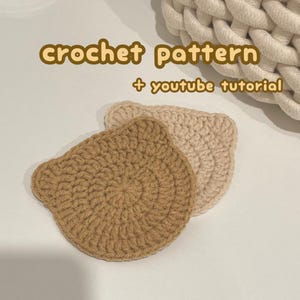 Puede incluir: Dos posavasos de ganchillo con forma de oso en beige y marrón claro. El texto "crochet pattern + youtube tutorial" se muestra en marrón. Los posavasos están hechos a mano y tienen un aspecto texturizado.