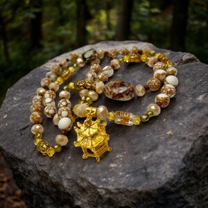 May include: A gold and white beaded necklace with a gold pagoda charm. The necklace is on a gray rock with a green background.