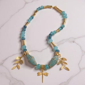 May include: A necklace with light and dark blue beads, linked by gold-coloured tubes. The necklace includes two large turquoise beads and gold leaf and dragonfly charms. The necklace is displayed on a white marble surface.