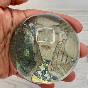 May include: A glass paperweight with a colorful image of a woman with flowers in her hair. The woman is wearing a yellow and blue patterned dress.
