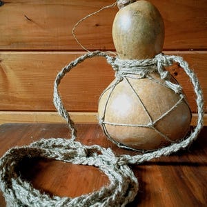 May include: A gourd-shaped container with a cork stopper, wrapped in a woven rope net and attached to a thick, braided rope handle. The gourd is a light brown color, and the rope is a natural, earthy tone.