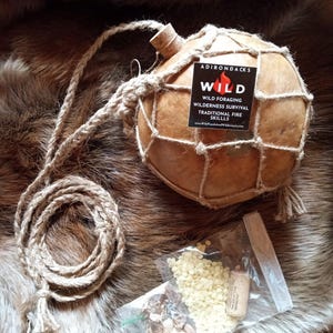 May include: A gourd water bottle with a woven net and rope handle, alongside a small bag of seeds and a cork. The bottle has a label that reads "Adirondacks Wild" with text about foraging and survival skills.