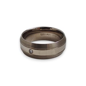 Può includere: Un anello in titanio grigio metallizzato caratterizzato da una fascia centrale lucida con una singola pietra preziosa rotonda e trasparente incastonata. L'interno dell'anello reca l'incisione "Pedio titanium". Profilo liscio e arrotondato.