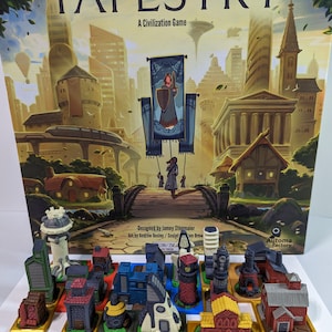 May include: A board game called Tapestry: A Civilization Game, with a colorful box featuring a cityscape and a banner with a woman holding a shield. The box is next to a set of miniature buildings, each on a gold base.
