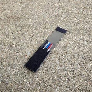 May include: A rectangular, black and gray accessory with a Velcro closure. Inside are four small, colored tubes: black, gray, red, and blue. The item is lying on a rough, gray concrete surface.