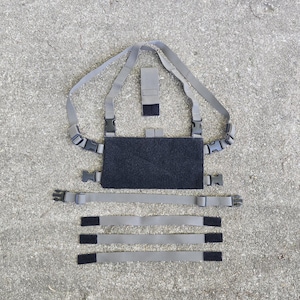 May include: A tactical chest rig in gray and black. The rig features a central black panel with adjustable straps, buckles, and additional modular attachment points. The design is laid out on a textured gray surface.
