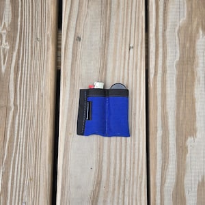 May include: A small, blue and black pouch with a flap closure, resting on a wooden surface. The pouch appears to be made of fabric and contains small items, including a white object and a black circular item.