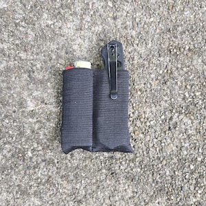 May include: A black nylon dual-pocket organizer pouch featuring a vertical stitched divider. One pocket contains a lighter with a red top, while the other pocket holds a folding pocket knife with a black handle and a silver metal pocket clip attached.