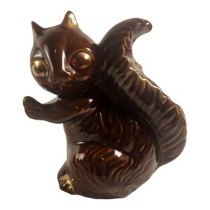 May include: A glossy dark brown ceramic squirrel figurine featuring gold-toned accents on the ears, eyes, and feet. The squirrel is depicted in a seated position with its large, bushy tail curved upward behind its back and one paw raised forward.