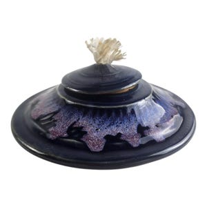 May include: A circular ceramic oil lamp with a glossy dark blue glaze. The center features a raised, tiered design with a radiating purple and white crystalline pattern. A white cotton wick protrudes from the top center of the lamp.