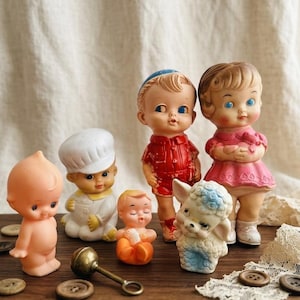 Pode incluir: Uma coleção de bonecos de borracha vintage, incluindo uma figura nua, um chef, um bebé em laranja, um rapaz com roupa vermelha, uma rapariga com vestido rosa e uma pequena figura de cordeiro, com um sino de latão e botões castanhos.
