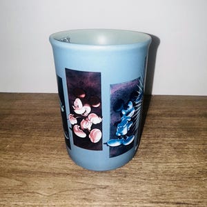 Peut inclure: Un gobelet en c&eacute;ramique bleu clair avec des images de Mickey Mouse. Le gobelet a une forme cylindrique et un bord &eacute;vas&eacute;. Les illustrations de Mickey Mouse sont sur un fond sombre.