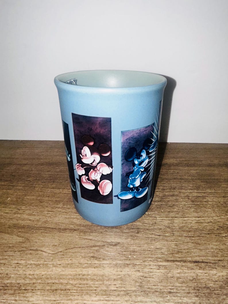 Peut inclure: Un gobelet en c&eacute;ramique bleu clair avec des images de Mickey Mouse. Le gobelet a une forme cylindrique et un bord &eacute;vas&eacute;. Les illustrations de Mickey Mouse sont sur un fond sombre.