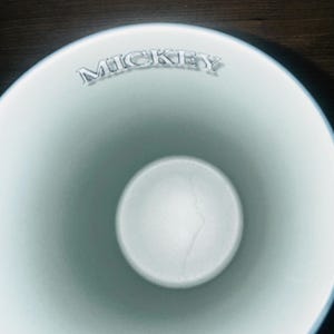 Peut inclure: Une tasse en c&eacute;ramique bleu clair avec le nom "MICKEY" en lettres argent&eacute;es sur le bord. L'int&eacute;rieur est d'une nuance de bleu plus claire, avec une base circulaire blanche. La tasse a une finition lisse et brillante.