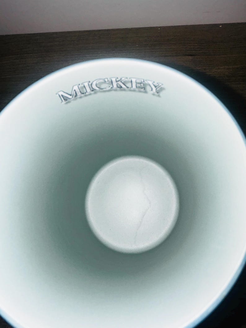 Peut inclure: Une tasse en c&eacute;ramique bleu clair avec le nom "MICKEY" en lettres argent&eacute;es sur le bord. L'int&eacute;rieur est d'une nuance de bleu plus claire, avec une base circulaire blanche. La tasse a une finition lisse et brillante.