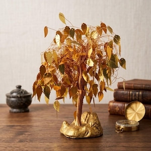 Könnte beinhalten: Eine dekorative Baumskulptur mit einem Stamm und Ästen aus gedrehtem Kupferdraht, verziert mit zahlreichen kleinen, metallisch goldfarbenen Blättern. Der Baum ist auf einem strukturierten, goldfarbenen Sockel befestigt.