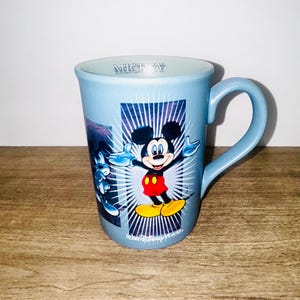 Peut inclure: Une tasse en c&eacute;ramique bleu clair repr&eacute;sentant Mickey Mouse les bras tendus. La tasse a une anse incurv&eacute;e et les mots "Mickey" sont imprim&eacute;s sur le bord. La tasse a un graphisme de Mickey Mouse sur fond bleu et blanc.