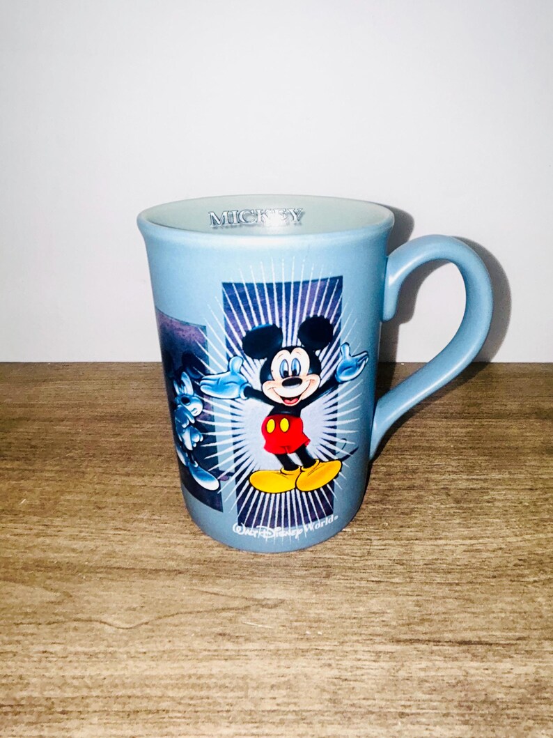 Peut inclure: Une tasse en c&eacute;ramique bleu clair repr&eacute;sentant Mickey Mouse les bras tendus. La tasse a une anse incurv&eacute;e et les mots "Mickey" sont imprim&eacute;s sur le bord. La tasse a un graphisme de Mickey Mouse sur fond bleu et blanc.