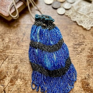 May include: A drawstring pouch covered in vibrant blue and black glass beads. The bag features horizontal bands of black beads alternating with sections of blue beads, finished with a long beaded fringe at the bottom and a braided cord handle.