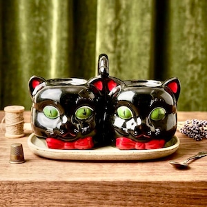 Può includere: Un set per condimenti in ceramica composto da due contenitori a forma di gatto nero con occhi verde brillante, orecchie interne rosse e papillon rossi. Le due teste di gatto sono unite da un manico centrale e poggiano su un vassoio ovale color crema.