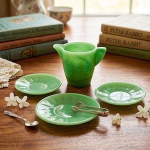 Peut inclure: Une crémière verte jade, trois soucoupes et une pince à sucre sont disposées sur une table en bois. Une cuillère en argent et des fleurs blanches ajoutent des détails. Des livres anciens sont empilés en arrière-plan, suggérant un cadre de thé.