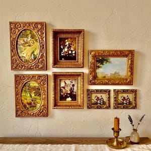 Puede incluir: Una colección de obras de arte vintage enmarcadas en marcos dorados ornamentados. Las obras de arte incluyen escenas de paisajes y naturalezas muertas florales. Los marcos son rectangulares y ovalados, creando una estética de pared de galería. Una vela y un jarrón de lavanda están sobre una superficie de madera.