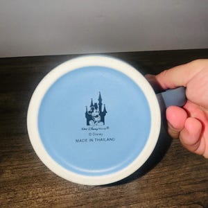 Peut inclure: Une tasse en c&eacute;ramique bleue et blanche avec un int&eacute;rieur bleu clair. Le fond de la tasse pr&eacute;sente un motif de ch&acirc;teau avec Mickey Mouse, le texte "Walt Disney World", "&copy; Disney" et "MADE IN THAILAND".