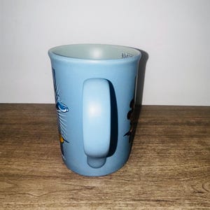 Peut inclure: Une tasse en c&eacute;ramique bleu clair avec une anse incurv&eacute;e. La tasse pr&eacute;sente des illustrations color&eacute;es de fleurs et d'un oiseau. Le mot "Mir" est visible sur le bord de la tasse. La tasse est pos&eacute;e sur un fond clair.
