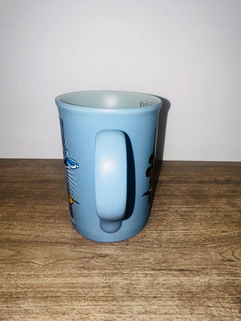 Peut inclure: Une tasse en c&eacute;ramique bleu clair avec une anse incurv&eacute;e. La tasse pr&eacute;sente des illustrations color&eacute;es de fleurs et d'un oiseau. Le mot "Mir" est visible sur le bord de la tasse. La tasse est pos&eacute;e sur un fond clair.