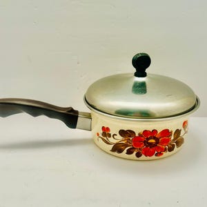 May include: Cream-colored saucepan with a stainless steel lid and a dark brown handle. The pan is decorated with red and brown floral designs. The handle is approximately 7 inches long.