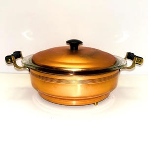 May include: Copper casserole dish with a glass lid and black handles. The dish has a copper finish and a gold trim.
