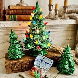 May include: Three green ceramic Christmas trees, with the largest one illuminated by multi-colored plastic bulbs. A small plastic bag labeled "LARGE SIZE TREE LIGHTS" containing extra replacement bulbs sits in front of the trees on a wooden surface.