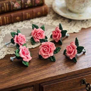 May include: A set of five drawer knobs shaped like pink roses with green leaves. Each knob features a detailed floral design with a metal screw extending from the back for attachment. The rose petals are textured and painted in shades of pink.