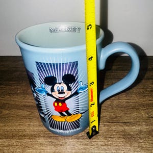 Peut inclure: Mug en c&eacute;ramique bleu clair avec Mickey Mouse les bras tendus. La tasse a une anse incurv&eacute;e et le mot "Mickey" est imprim&eacute; sur le bord. La tasse mesure environ 10 cm de haut.