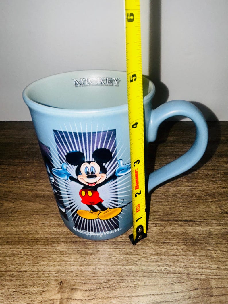Peut inclure: Mug en c&eacute;ramique bleu clair avec Mickey Mouse les bras tendus. La tasse a une anse incurv&eacute;e et le mot "Mickey" est imprim&eacute; sur le bord. La tasse mesure environ 10 cm de haut.