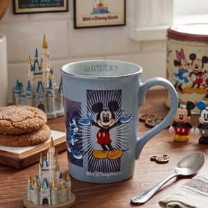 Peut inclure: Une tasse en c&eacute;ramique bleu clair avec Mickey Mouse et le mot "Mickey" sur le bord. La tasse est entour&eacute;e d'objets sur le th&egrave;me de Disney, notamment des ch&acirc;teaux miniatures et des biscuits, sur une surface en bois.