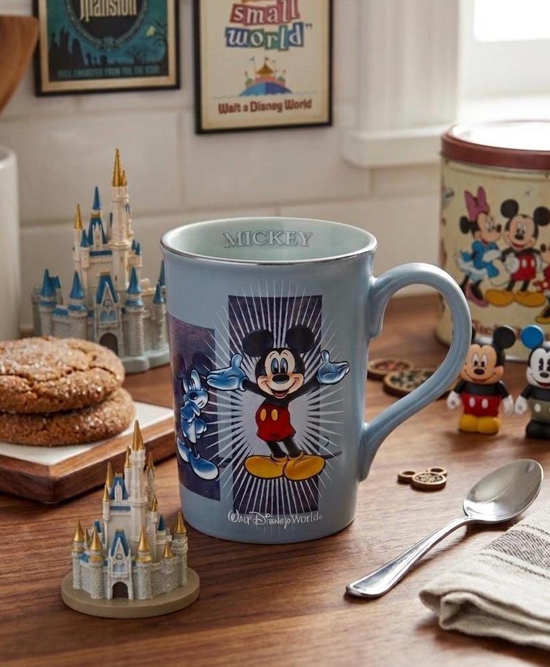 Peut inclure: Une tasse en c&eacute;ramique bleu clair avec Mickey Mouse et le mot "Mickey" sur le bord. La tasse est entour&eacute;e d'objets sur le th&egrave;me de Disney, notamment des ch&acirc;teaux miniatures et des biscuits, sur une surface en bois.