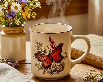 May include: A cream-colored mug with a butterfly design, filled with a steaming beverage. The butterfly is red and black, surrounded by floral elements. A small vase of wildflowers sits nearby on a wooden surface.