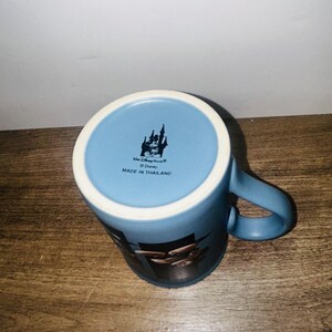 Peut inclure: Mug en c&eacute;ramique bleue avec un bord blanc et une anse incurv&eacute;e. Le mug pr&eacute;sente un motif Disney World et le texte "Made in Thailand". Le mug est pos&eacute; sur une surface en bois.