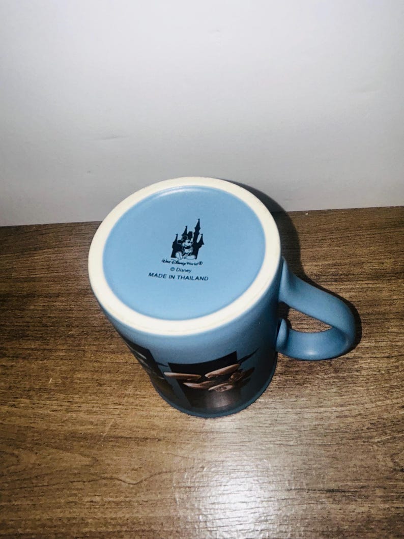Peut inclure: Mug en c&eacute;ramique bleue avec un bord blanc et une anse incurv&eacute;e. Le mug pr&eacute;sente un motif Disney World et le texte "Made in Thailand". Le mug est pos&eacute; sur une surface en bois.