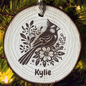 Handmade rustic sliced wood ornament featuring a floral bird design, with delicate flowers and leaves surrounding the bird. The ornament is custom personalized with a name, and the design is laser engraved for a unique, thoughtful home décor gift.