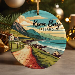 May include: A round ceramic ornament with a scenic image of Keem Bay, Ireland. The ornament features a coastal landscape with a winding road, green hills, and a blue ocean. The text "Keem Bay, Ireland" is printed on the ornament.