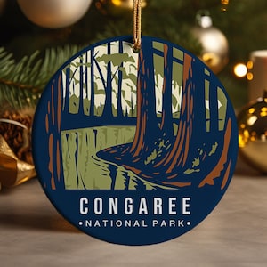 May include: Round Christmas ornament with a forest scene illustration. The ornament is dark blue with a gold string for hanging. The illustration features trees, a river, and the text "CONGAREE NATIONAL PARK."